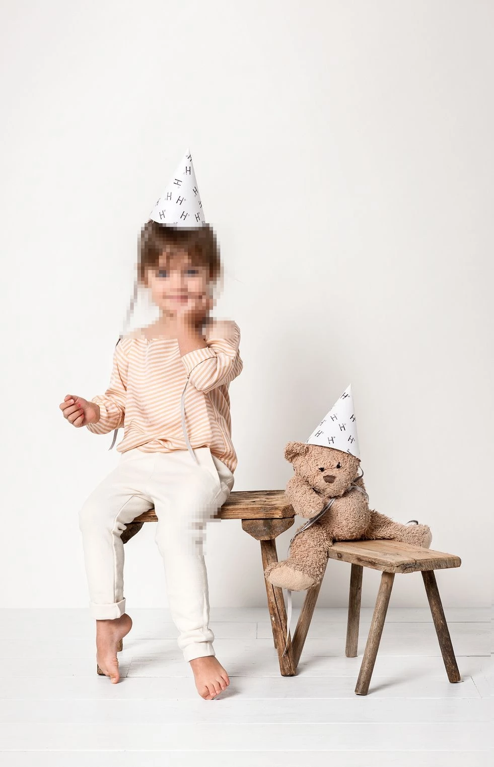 Little girl in party hat with teddy bear on rustic stools in minimalist studio