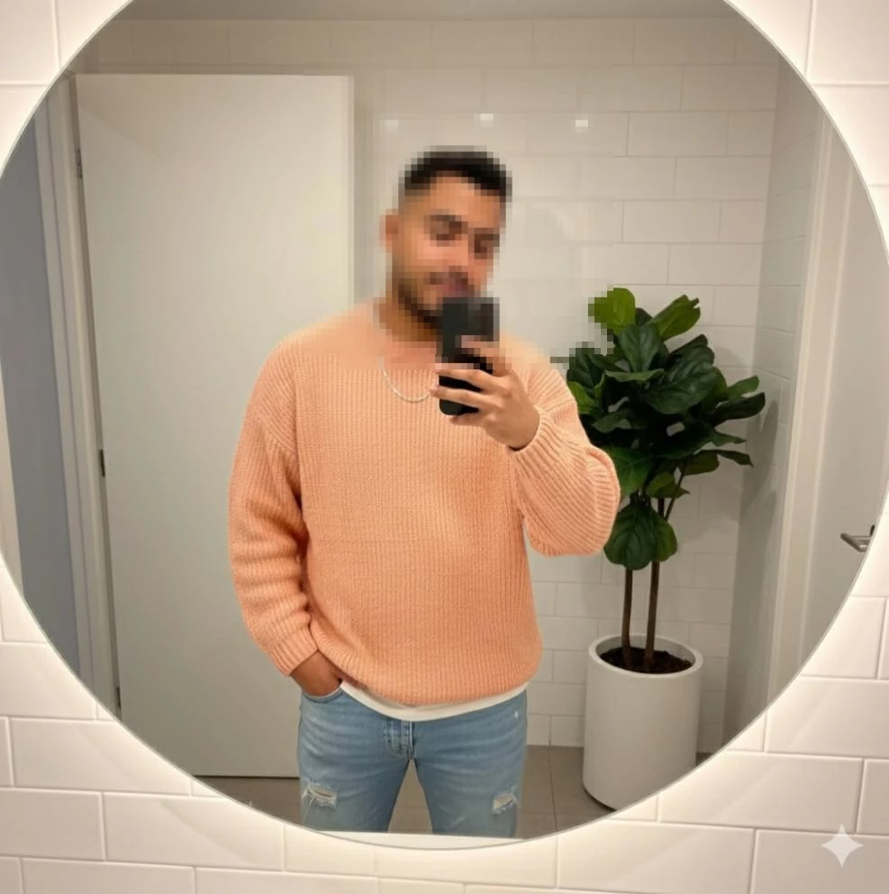 Man taking mirror selfie wearing peach sweater and light blue jeans in modern bathroom