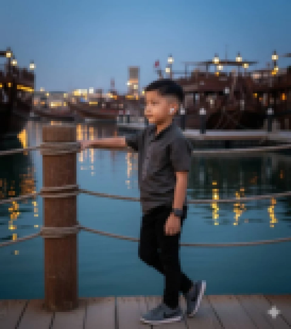 Young boy standing on a dock at sunset with boats and lights in the background