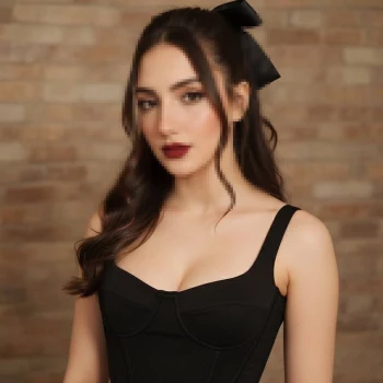 Elegant woman in black dress with wavy hair and bow posing against brick wall