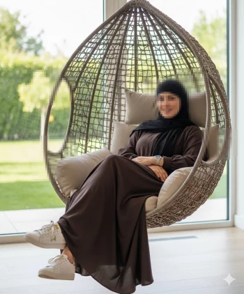 Elegant woman in brown abaya sitting on hanging chair by large window
