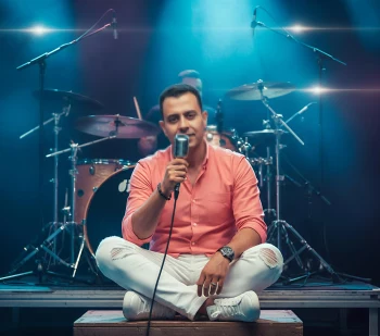 Man in salmon pink shirt singing on stage with live band and blue lights