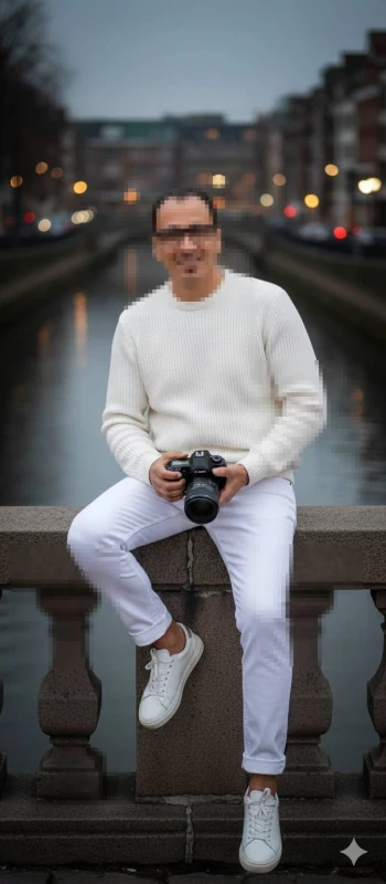 Man in white outfit holding camera sitting by city canal at dusk