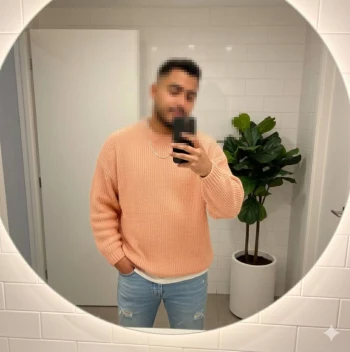 Man taking mirror selfie wearing peach sweater and light blue jeans in modern bathroom
