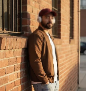 Man wearing brown jacket and burgundy cap leaning on brick wall with headphones
