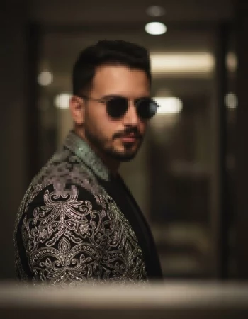 Man wearing embroidered jacket and sunglasses posing indoors with soft lighting
