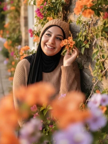 Smiling woman in black hijab and brown sweater surrounded by colorful flowers
