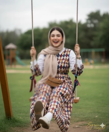 Smiling woman in patterned dress and beige hijab enjoying swing in green park