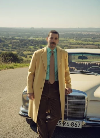 Stylish man in yellow coat standing beside vintage car on scenic countryside road